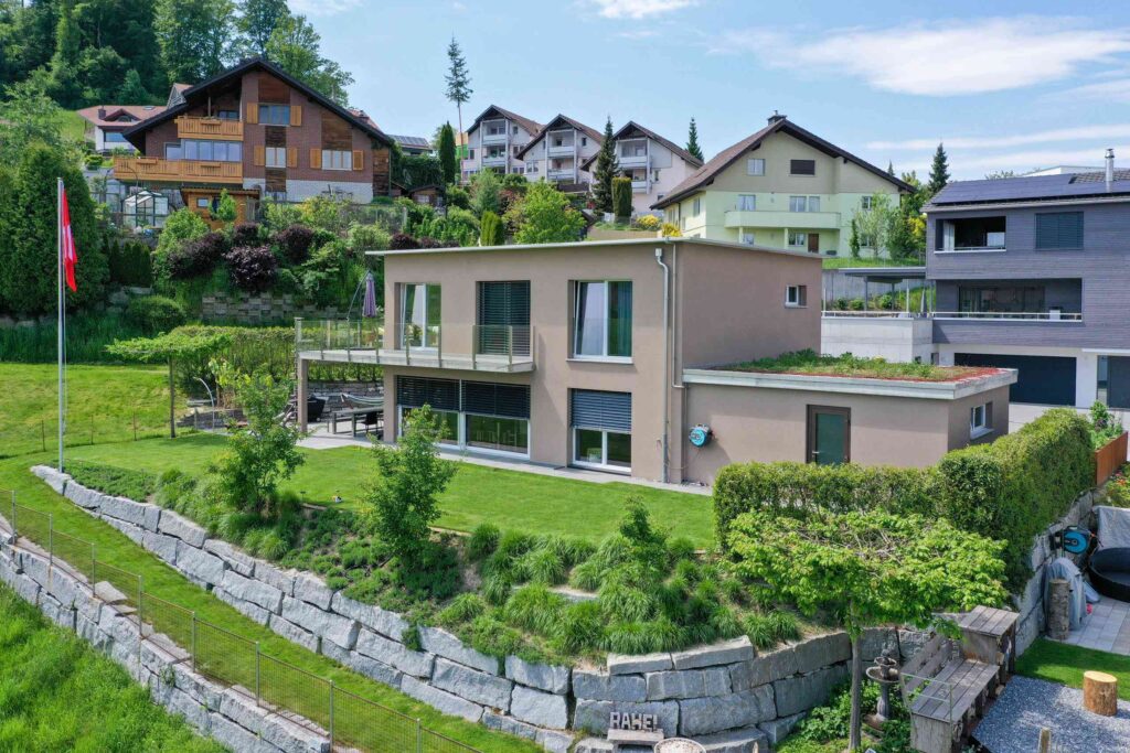Modernes zweistöckiges Haus mit großen Fenstern und einer Terrasse, umgeben von einem gepflegten Rasen und Garten. Mehrere Nachbarhäuser und eine Bergkulisse sind sichtbar.
