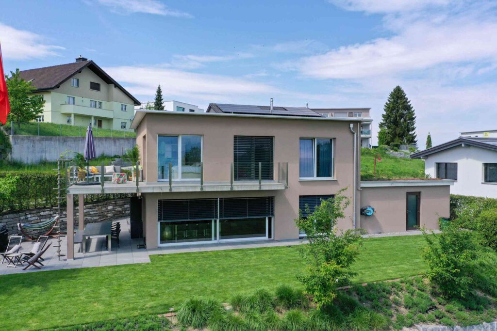 Modernes zweistöckiges Haus mit großen Fenstern, einer Solaranlage auf dem Dach und einer Terrasse mit Gartenmöbeln, umgeben von gepflegten Grünanlagen und in einer Vorstadtgegend gelegen.