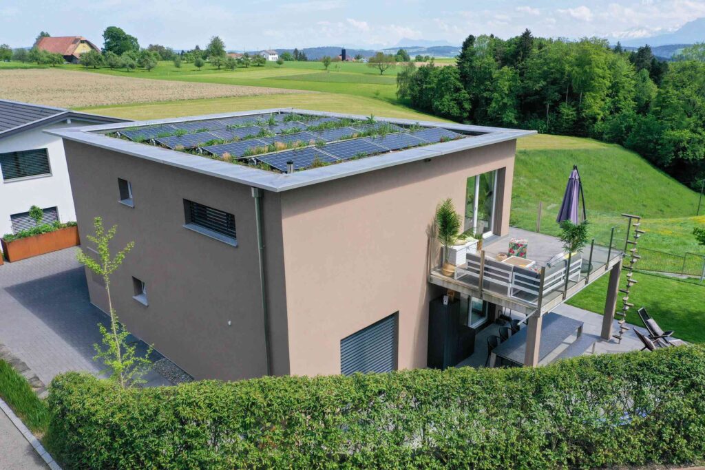 Ein modernes, würfelförmiges Haus mit Dachgarten und Sonnenkollektoren, gelegen in einer grünen, ländlichen Landschaft mit Hügeln im Hintergrund.