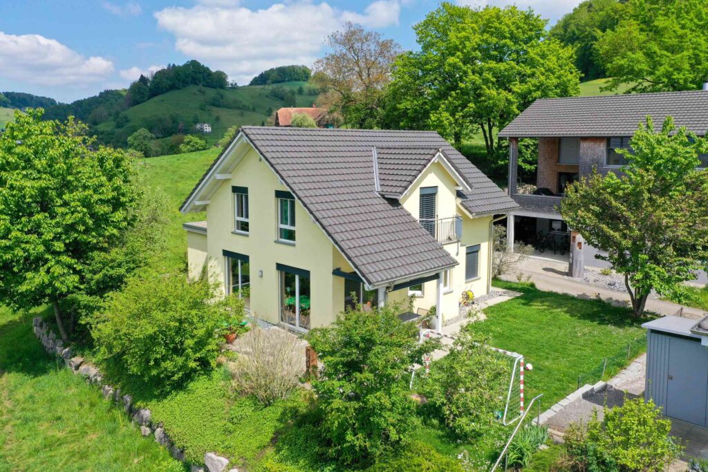 Ein gelbes Haus mit grauem Dach ist von üppigem Grün und Bäumen umgeben und in eine hügelige, ländliche Landschaft eingebettet.