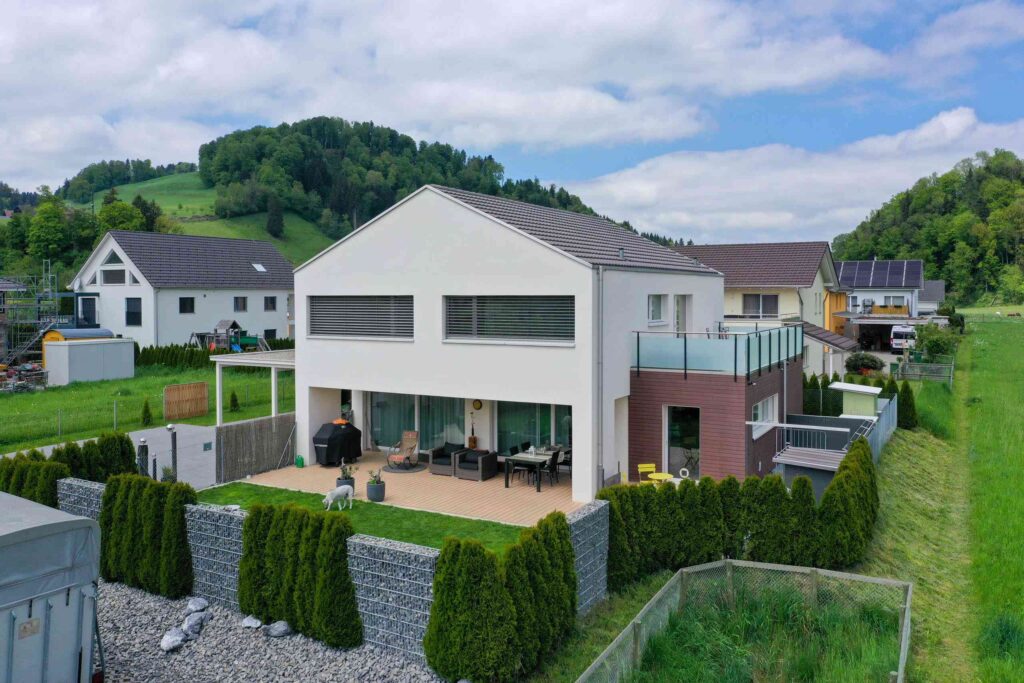 Modernes zweistöckiges Haus mit großen Fenstern, einer Terrasse und einem Balkon, in einer grünen Vorstadtgegend mit umgebenden Hügeln und Nachbarhäusern.