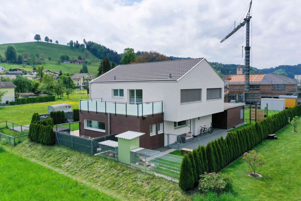 Ein modernes zweistöckiges Haus mit Glasbalkon, gelegen in einem grasbewachsenen Wohngebiet in der Nähe eines Hügels, im Hintergrund ist ein Baukran zu sehen.