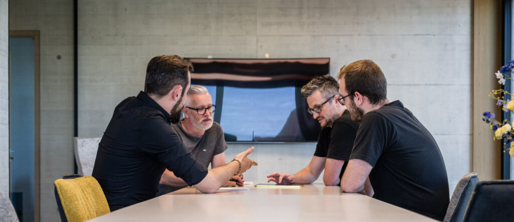 Vier Männer sitzen in einer modernen Büroumgebung um einen Konferenztisch und diskutieren. Ein Mann gestikuliert, während die anderen zuhören.