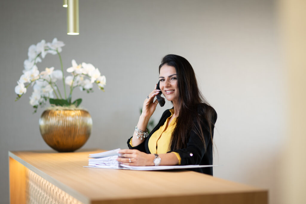Eine Frau steht an einem Empfangstresen, hält Papiere in der Hand und telefoniert. Im Hintergrund ist eine goldene Vase mit weißen Orchideen zu sehen.