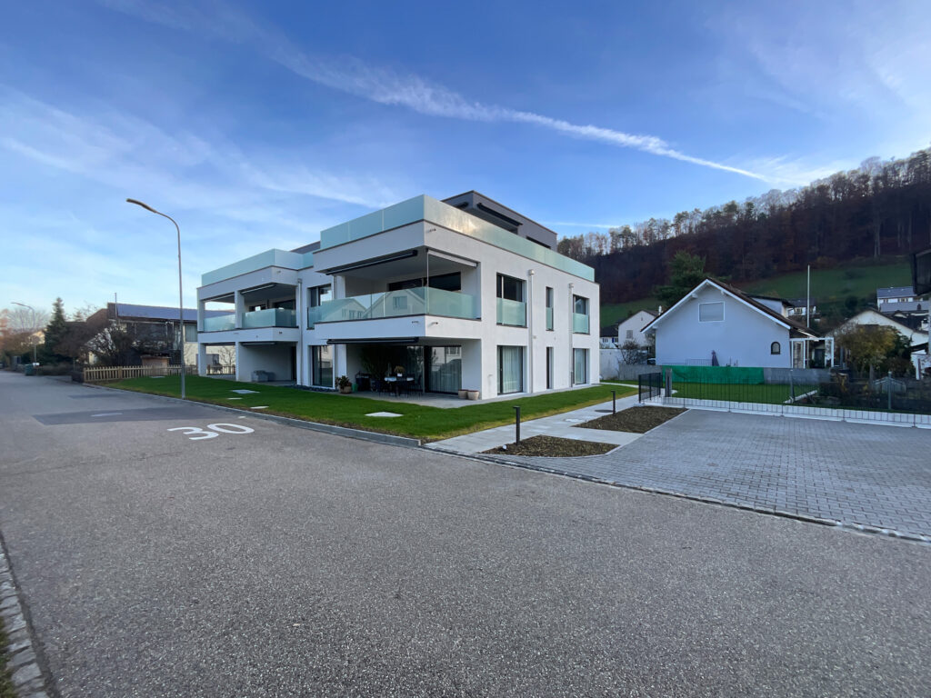 Modernes zweistöckiges weißes Gebäude mit großen Glasbalkonen auf einem Eckgrundstück an einer Vorortstraße mit Geschwindigkeitsbegrenzungsmarkierung und nahe gelegenen Häusern.